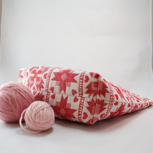May include: A white and red patterned fabric tote bag with two pink yarn balls in front of it.