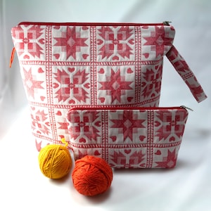 May include: Two red and white patterned fabric zippered pouches. The larger pouch has a handle. The smaller pouch is sitting inside the larger pouch. Two balls of yarn are in front of the pouches.