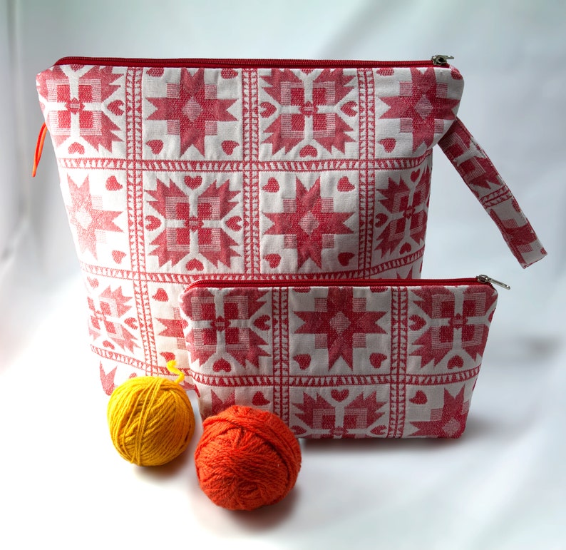 May include: Two red and white patterned fabric zippered pouches. The larger pouch has a handle. The smaller pouch is sitting inside the larger pouch. Two balls of yarn are in front of the pouches.