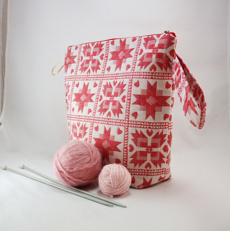 May include: A white and red patterned fabric project bag with a zipper closure and a handle. Two pink yarn balls and two knitting needles are in front of the bag.