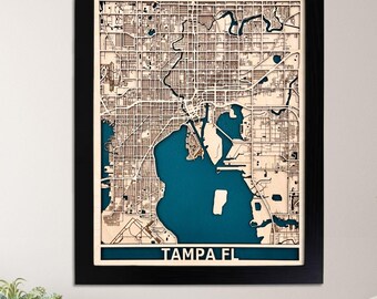 May include: A wooden map of Tampa, Florida, in a black frame. The map is laser cut and features a blue water body. The text "TAMPA FL" is printed below the map. The image also shows three icons: a green leaf with a hand, a map of the United States, and a thumbs up with five stars.