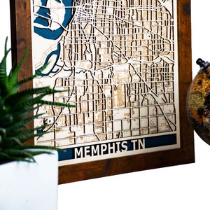May include: A wooden framed map of Memphis, Tennessee with a blue and white color scheme. The map is laser cut and shows the streets of the city. The frame is brown wood.