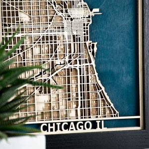May include: A framed, layered wooden map of Chicago, Illinois, with a teal blue background. The city's grid-like street layout is intricately cut from light-coloured wood. The frame is black, and the text "CHICAGO IL" is visible at the bottom.