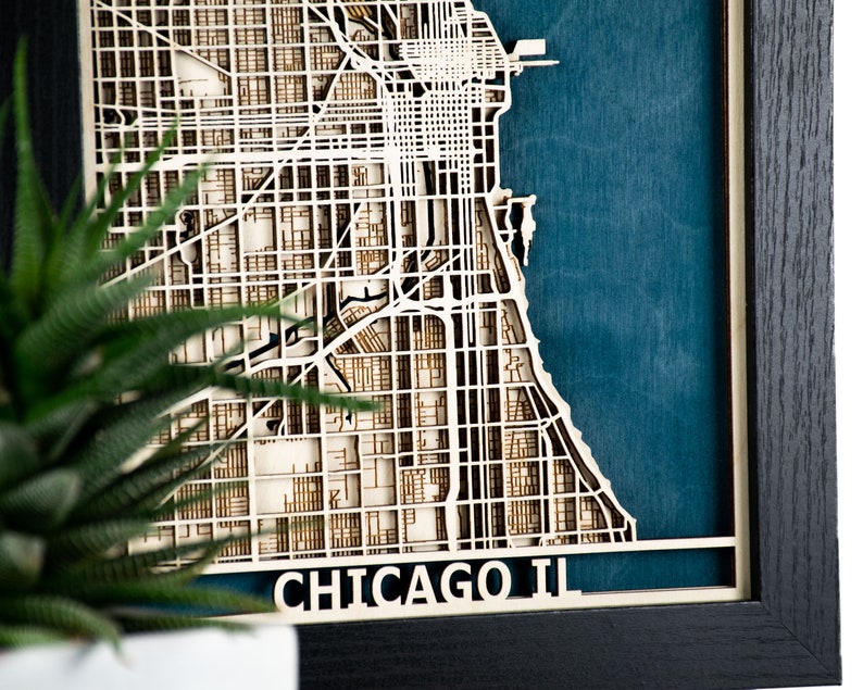 May include: A framed, layered wooden map of Chicago, Illinois, with a teal blue background. The city's grid-like street layout is intricately cut from light-coloured wood. The frame is black, and the text "CHICAGO IL" is visible at the bottom.