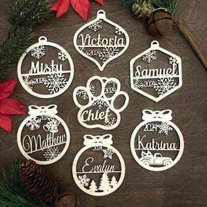 Op de afbeelding: Zeven gepersonaliseerde houten kerstversieringen met sneeuwvlokontwerpen. De ornamenten hebben de namen "Victoria", "Misty", "Samuel", "Chief", "Matthew", "Katrina" en "Evalyn" met het jaar "2024" op elk.
