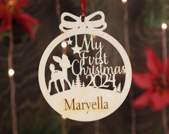 May include: A wooden Christmas ornament shaped like a bauble with a deer silhouette, the text "My First Christmas 2024" and the name "Maryella".