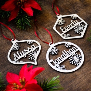 Op de afbeelding: Drie witte houten kerstversieringen met sneeuwvlokontwerpen en gepersonaliseerde namen: Alyssa, Everly en Jonathan. De versieringen hangen aan een rood lint.