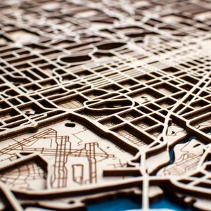 May include: A detailed wooden map, laser-cut to show a city's street layout. The map has precise cuts defining roads, buildings, and waterways, with a natural wood grain finish. A blue water feature adds contrast to the light brown wood, creating a striking visual.