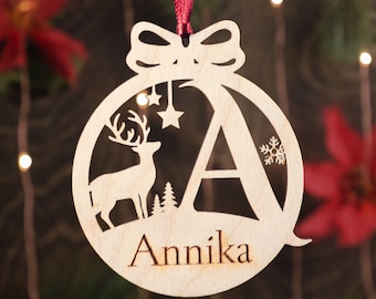 May include: A wooden Christmas ornament shaped like a ball with a deer, stars, and a snowflake cut out. The ornament is personalized with the letter "A" and the name "Annika".