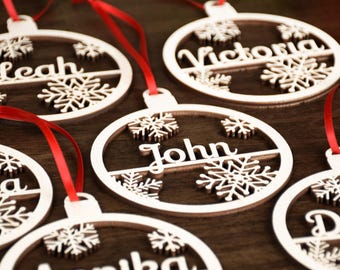 May include: Five white wooden Christmas ornaments with snowflake designs and personalized names: Leah, Victoria, John, Annika, and D. Each ornament has a red ribbon attached.