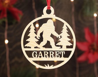 May include: A wooden Christmas ornament with a Bigfoot silhouette walking between two trees. The ornament is round with the name "GARRET" cut out at the bottom. It hangs from a red ribbon.