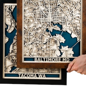 May include: Two framed laser-cut maps of cities. The top frame is brown wood and features a map of Baltimore, MD. The bottom frame is black and features a map of Tacoma, WA. Both maps have a blue water feature.