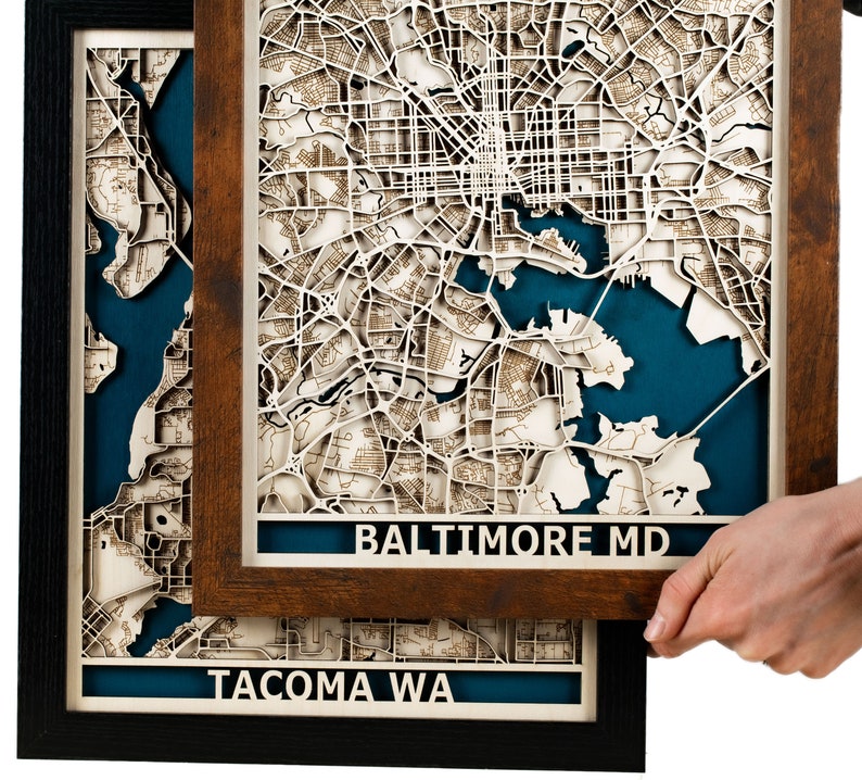 May include: Two framed laser-cut maps of cities. The top frame is brown wood and features a map of Baltimore, MD. The bottom frame is black and features a map of Tacoma, WA. Both maps have a blue water feature.