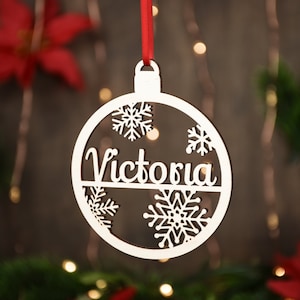 Op de afbeelding: Witte houten kerstbal ornament met de naam "Victoria" uitgesneden en sneeuwvlokken rond de rand.