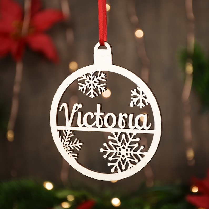 Op de afbeelding: Witte houten kerstbal ornament met de naam "Victoria" uitgesneden en sneeuwvlokken rond de rand.