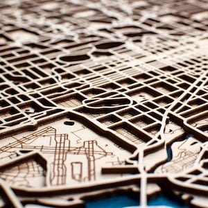 May include: A detailed wooden map of a city, with intricate laser-cut lines representing streets and waterways. The map is made of light brown wood and has a raised, three-dimensional effect.