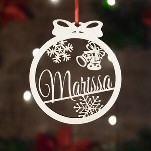 Op de afbeelding: Witte houten kerstbal met een strik aan de bovenkant. De kerstbal heeft een sneeuwvlokkenpatroon, bellen en de naam "Marissa" uitgesneden.