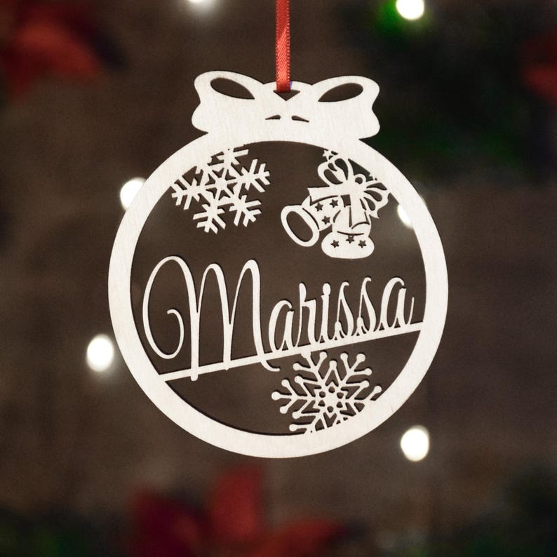 Op de afbeelding: Witte houten kerstbal met een strik aan de bovenkant. De kerstbal heeft een sneeuwvlokkenpatroon, bellen en de naam "Marissa" uitgesneden.