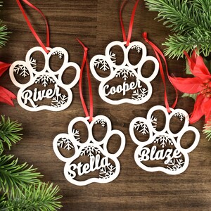 Op de afbeelding: Vier witte houten pootvormige ornamenten met sneeuwvlokmotieven en gepersonaliseerde namen: River, Cooper, Stella en Blaze. De ornamenten hangen aan rode linten.