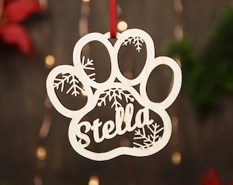 May include: A white wooden paw print ornament with the name "Stella" cut out and snowflake designs. The ornament is hanging from a red ribbon.