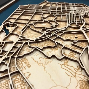 May include: A detailed wooden map of a city, possibly San Francisco, with intricate laser-cut streets and topographical features. The map is made of light-coloured wood with darker wood outlining the streets, creating a 3D effect. The map is mounted on a blue background.