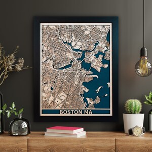May include: A framed wooden map of Boston, Massachusetts, with detailed laser-cut features. The map shows a dark blue background for the water, and the city's streets and buildings are in light brown wood. The text "BOSTON MA" is at the bottom of the frame.
