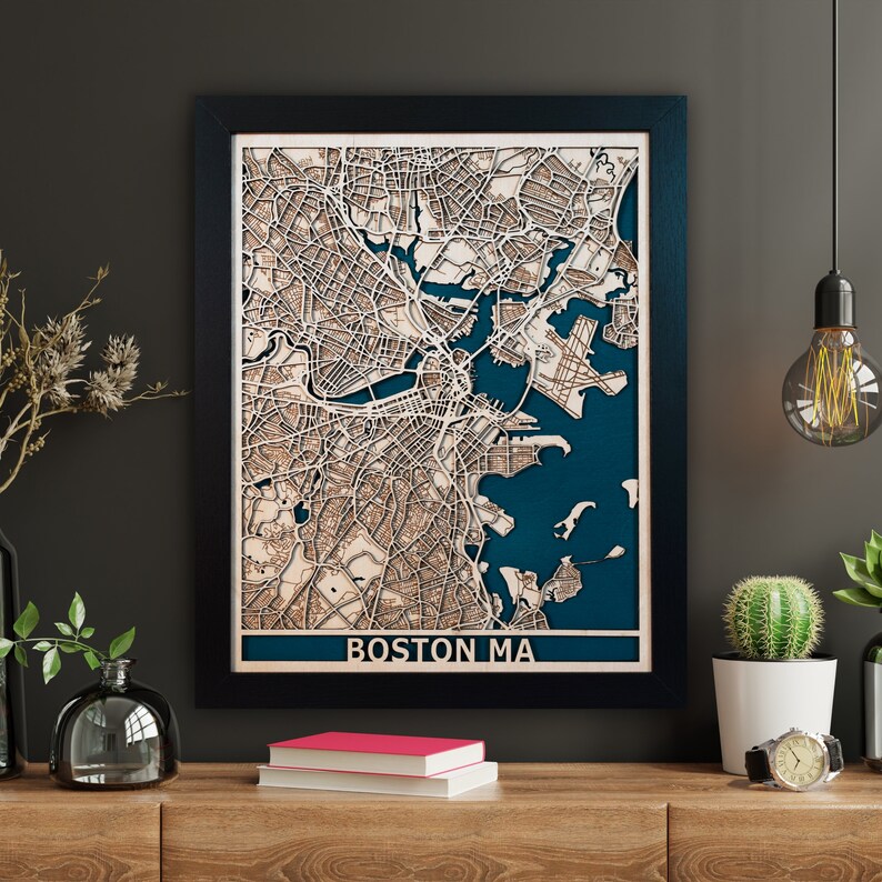 May include: A framed wooden map of Boston, Massachusetts, with detailed laser-cut features. The map shows a dark blue background for the water, and the city's streets and buildings are in light brown wood. The text "BOSTON MA" is at the bottom of the frame.