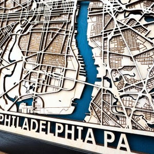 May include: A wooden map of Philadelphia, Pennsylvania, with a blue water feature representing the Delaware River. The map is laser cut and features detailed street outlines. The text "PHILADELPHIA PA" is visible at the bottom of the map.