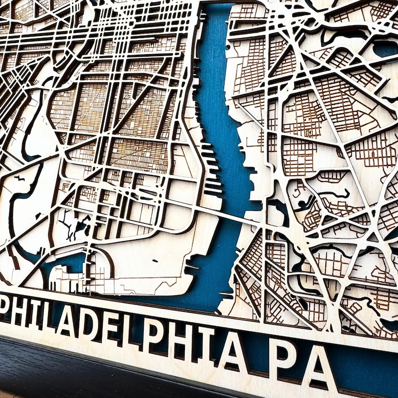 May include: A wooden map of Philadelphia, Pennsylvania, with a blue water feature representing the Delaware River. The map is laser cut and features detailed street outlines. The text "PHILADELPHIA PA" is visible at the bottom of the map.