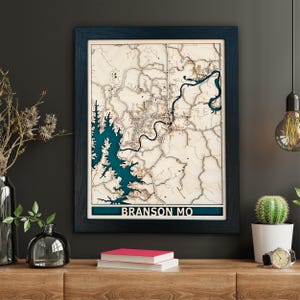 May include: A wooden 3D map of Branson, Missouri, in a dark blue frame. The map features a blue lake and brown land. The text "BRANSON MO" is printed on the bottom of the map.