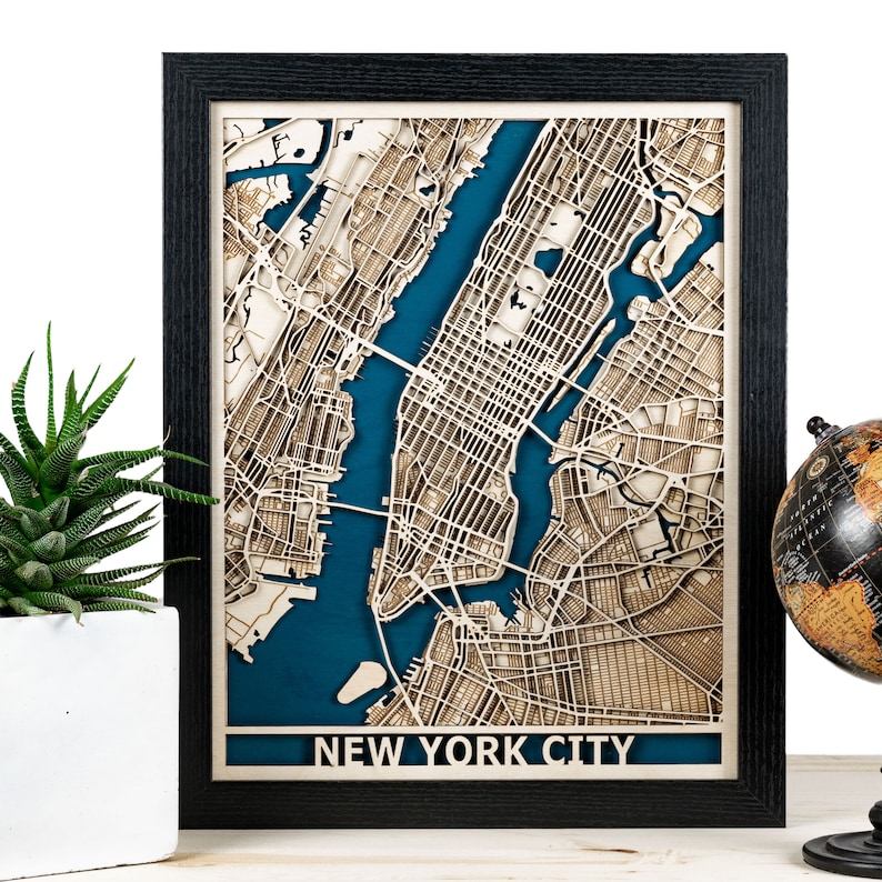 May include: A framed wooden map of New York City, with a detailed laser-cut design. The map shows the city's layout with a blue background representing the water. The frame is black, and the text "NEW YORK CITY" is displayed at the bottom.