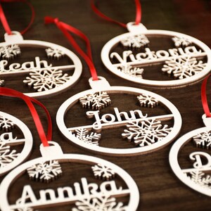 Op de afbeelding: Zes houten kerstversieringen met sneeuwvlokontwerpen en gepersonaliseerde namen: Leah, Victoria, John, Annika en twee anderen. De versieringen hangen aan een rood lint.