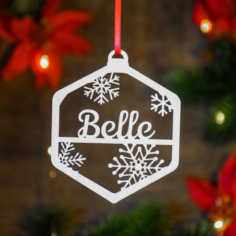 Op de afbeelding: Wit houten kerstversiering in de vorm van een hexagon met sneeuwvlokken en de naam "Belle" in een decoratief lettertype.