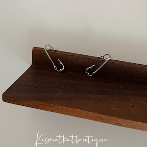 Peut inclure: Deux boucles d'oreilles en forme d'épingle de sûreté argentées sur une surface en bois. Les boucles d'oreilles sont simples et minimalistes.