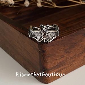 Puede incluir: Un anillo de metal plateado con un diseño detallado de murciélago con las alas extendidas. El murciélago está centrado en la banda, mostrando alas texturizadas y una cabeza pequeña. El texto Kismetkatboutique es visible en letras blancas.