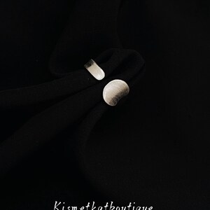 May include: A silver-toned open ring featuring a flat, rounded circular element on one end and a rectangular bar on the other, resting on black fabric. The text Kismetkatboutique is printed in white at the bottom of the image.