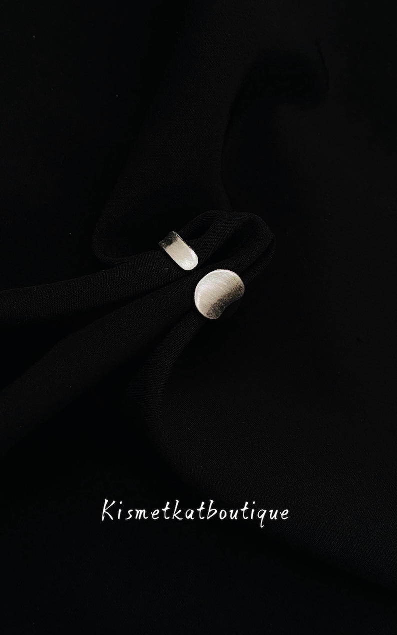 May include: A silver-toned open ring featuring a flat, rounded circular element on one end and a rectangular bar on the other, resting on black fabric. The text Kismetkatboutique is printed in white at the bottom of the image.