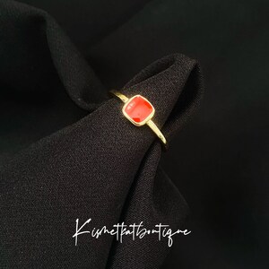 May include: A gold-colored ring with a square, red gemstone. The ring is set against a black background. The text "Kismetkatboutique" is written in cursive on the black fabric.