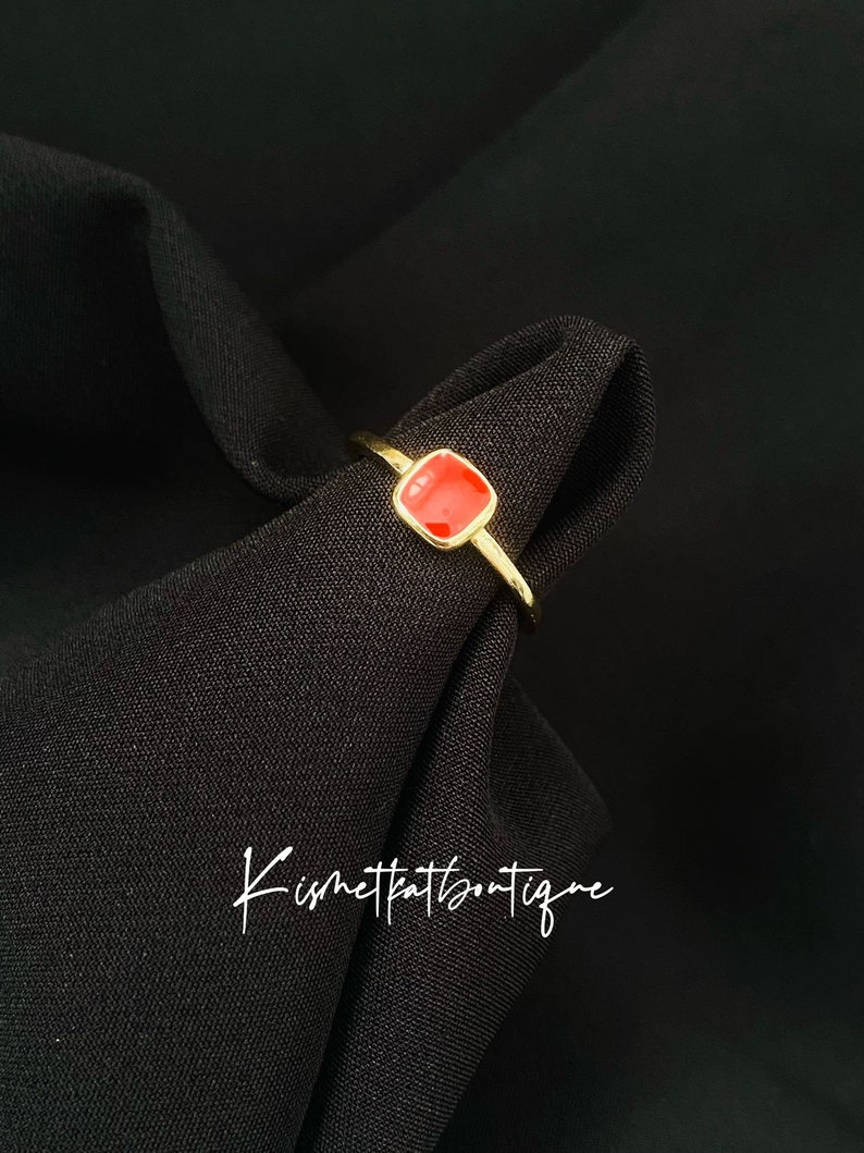May include: A gold-colored ring with a square, red gemstone. The ring is set against a black background. The text "Kismetkatboutique" is written in cursive on the black fabric.