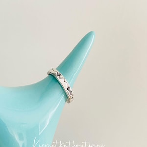 May include: A silver ring with a geometric pattern on a light blue ring holder. The ring is on a light blue surface with the text "kismetkatboutique" in black.