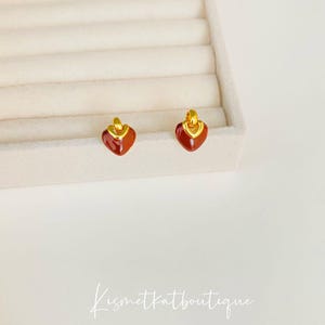 Może przedstawiać: Para złotych kolczyków w kształcie serca z czerwoną emalią. Kolczyki są prezentowane w białym pudełku na biżuterię.