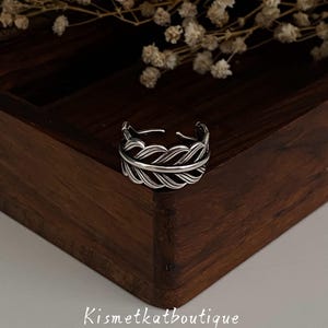 May include: A silver adjustable ring with a feather design. The ring has an open base and a detailed pattern of overlapping lines. It is displayed on a dark wooden surface, with dried flowers in the background. The text "Kismetkatboutique" is visible.