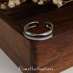 May include: A silver-toned open ring featuring a double-band design. One band is smooth and polished, while the other is thinner and encrusted with small, clear, sparkling stones. The interior of the band is stamped with the text 925.
