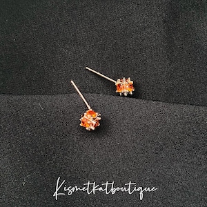 Puede incluir: Un par de pendientes de botón de oro con pequeñas piedras de circonita cúbica de color naranja. Los pendientes están sobre un fondo negro.