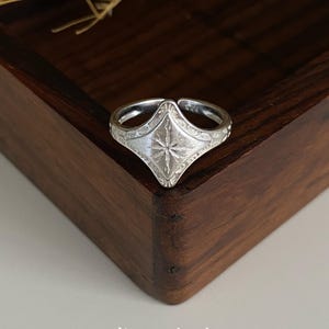 Könnte beinhalten: Ein silberner Ring mit einer rautenförmigen Vorderseite, die ein eingraviertes Sternenmuster und filigrane Details entlang der Ringschiene aufweist. Der Ring ist hinten offen und der Text Kismetkatboutique steht darunter auf der Oberfläche.