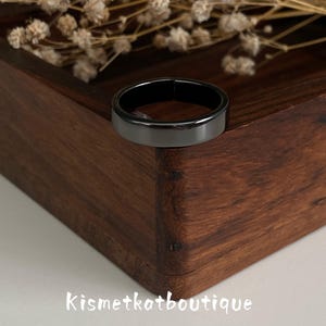 May include: A smooth, polished black band ring with a flat profile and a glossy finish. The ring is positioned on the corner of a dark brown wooden box. The text Kismetkatboutique is printed in white letters on the lower portion of the image.