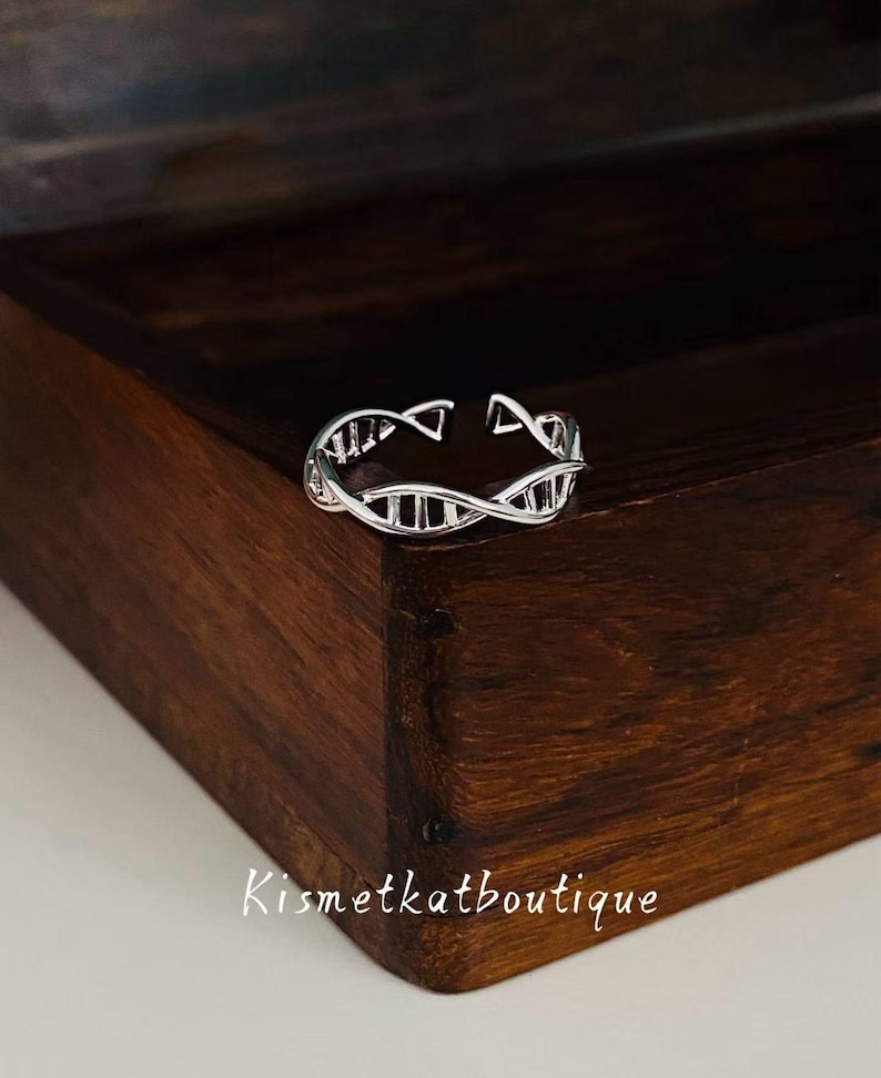 May include: A silver ring featuring a DNA double helix design. The ring is open at the top and has a detailed pattern of connected lines and shapes. The ring is displayed on a dark wooden surface. The text "Kismetkatboutique" is visible.