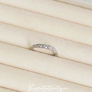Op de afbeelding: Een zilverkleurige ring met een gevlochten ontwerp, bezet met kleine, heldere edelstenen. De ring wordt gepresenteerd op een lichtbeige, getextureerd oppervlak. De tekst "Kismetkatboutique" is in cursief geschreven.
