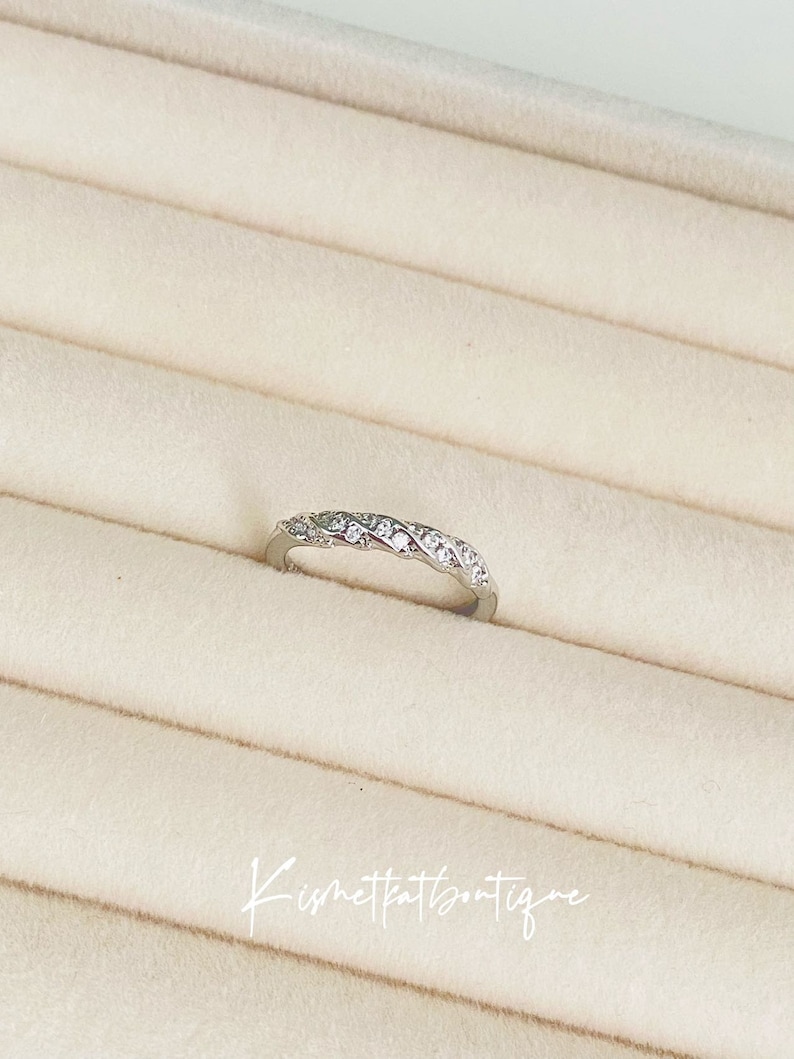 Op de afbeelding: Een zilverkleurige ring met een gevlochten ontwerp, bezet met kleine, heldere edelstenen. De ring wordt gepresenteerd op een lichtbeige, getextureerd oppervlak. De tekst "Kismetkatboutique" is in cursief geschreven.