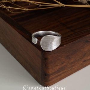 Könnte beinhalten: Ein silberfarbener offener Ring mit einem glatten, abgerundeten, flachen kreisförmigen Mittelpunkt mit gebürsteter Metalloberfläche. Die Ringschiene ist verstellbar und weist eine dezente, matte Textur auf. Der Text Kismetkatboutique ist unten sichtbar.